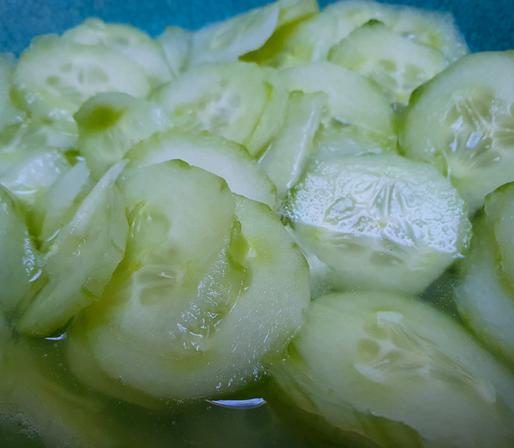Nahaufnahme eines Gurkensalats in einer türkisfarbenen Schale. Der Salat besteht aus dünn geschnittenen, hellgrünen Gurkenscheiben, die in einer klaren Marinade liegen. Die Gurkenscheiben sind teilweise überlappend angeordnet und wirken frisch, glänzend und saftig.