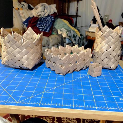 A lineup of 4 handmade baskets (one very tiny) on a small table.
The baskets are all made from strips cut from brown paper bags, folded and woven in various ways.