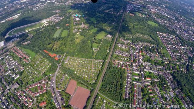 An aerial view towards Gelsenkirchen Feldmark. A railway track is going through the image. There are lobing quaters inside green oasis with trees all around. The harness racing track of Gelsenkirchen can be seen at the top left.