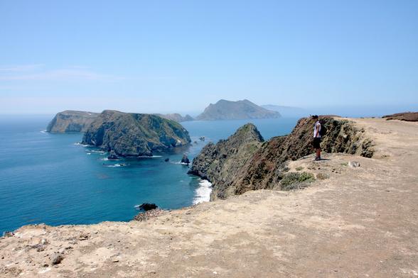 A teenage boy stands at the top of a dusty dry cliff overlooking the ocean. A chain of small rocky islands leads off into the background.