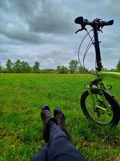 Grüne Wiese. Im Vordergrund zwei Beine mit Fahrradschuhen und rechts der Vorderteil eines Faltrades