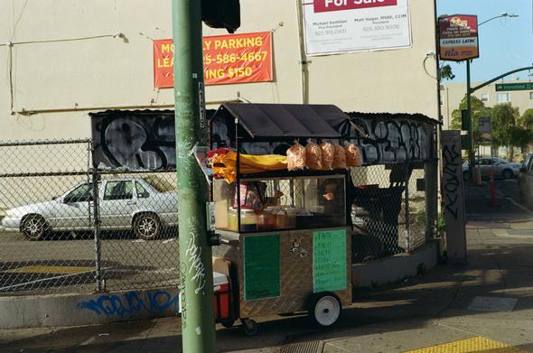 A food cart sells fruit on a street corner. Behind is a Volvo S70 behind a fence.
