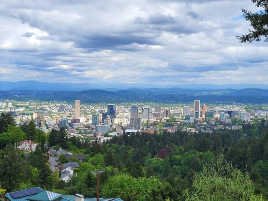 A long distance photo of the Portland skyline taken from Pittock Mansion. I'm the foreground are thick green trees on a downward slope. I'm the middle ground is the skyline of Portland, with a few skyscrapers and a variety of small buildings. In the distance is a line of mountains, although Mount Hood is obscured by clouds. All of this is under a bluish grey sky of puffy clouds.