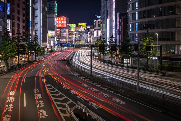 Long exposure shot on a buzzy main road of Shinjuku in Tokyo from a bridge over the road, at night. Only the light trails of the cars are visible on the road in the foreground due to the long exposure. Skyscrappers and buildings covered in colourful neons are recognisable in the background.