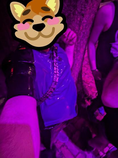 Photo of me in purple and black latex, wearing a shoulder pack, lit by some really queer purple lighting