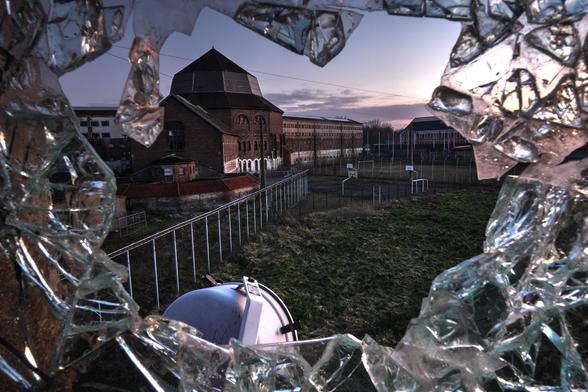 Foto eines Sonnenuntergangs, aufgenommen durch ein Loch einer eine eingeschlagenen Mehrschicht-Laminats-Sicherheitsglas-Scheibe. Blick auf das Zentralgebäude eines vikorianischen Gefängnisgebäudes in dunklem Backstein, überwucherter Bolzplatz im Vordergrund. Am unteren Bildrand Suchscheinwerfer des Wachturms. Über Kopf Stahlseile gegen Hubschrauberlandungen.