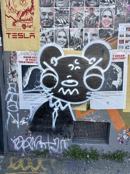 A graffiti drawing of a cute weasel-inspired character on a wall filled with posters and other graffiti. It's a two-color "throwie" with a black background and white detail lines. It's by an artist named Weasl and has a distinct style.
