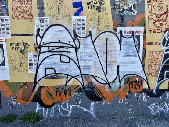 A graffiti drawing of a viking-inspired character on a wall filled with posters and other graffiti. It's a two-color "throwie" with a white background and black detail lines. It's by an artist named Ola The Barbarian and has "Ola" written under the character in a hard to read graffiti style.