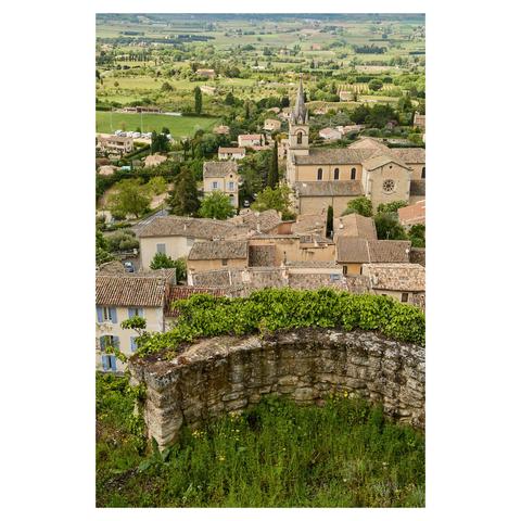 View over the rooftops of a village, taken from an elevated viewpoint. The church is in the center of the image. In the foreground, a curved and partly overgrown wall frames the image.