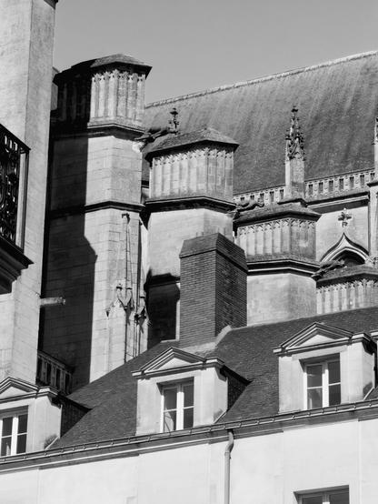 Cette photographie en noir et blanc représente une partie d'un vieux bâtiment d'architecture gothique. L'image se concentre sur des éléments d'architectures comme des pointes d'arches et des détails de gravures. Le bâtiment se trouve derrière la toiture d'une maison d'architecture beaucoup plus récente qui couvre le bas de l'image.