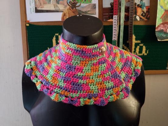 Cuello con pechera tejido a crochet en colores vibrantes (verde, rosa, naranja, morado), con textura de puntos cerrados y borde ondulado. Está abrochado con un botón dorado. El cuello está sobre un torso de maniquíe negro.