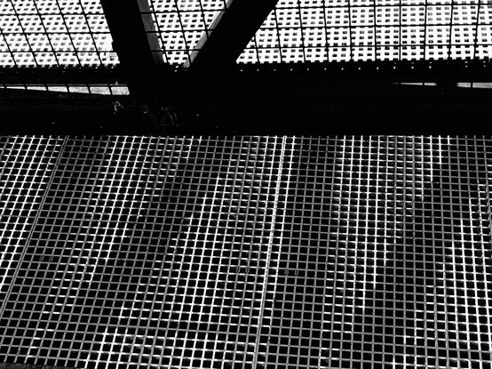 The photo is divided roughly into two parts. One part consisting of the lower 2/3 of the frame shows a metal grate with sunlight reflecting off the structure. Dark shadows angle across the grate cast by a fence. The fence separates that grate from another grate, which is very light. The overall effect is of many different sizes of squares.
