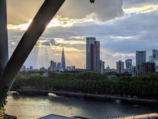 A view from South-East of the centre of London looking back at the main skyline, with the Shard central, god rays pouring down fron between some clouds behind it, the 'Walkie Talkie' visible off to the right, and the ugly new Founding building at Canada Water taking up sky in front of it all. A dock sprawls below in the foreground.