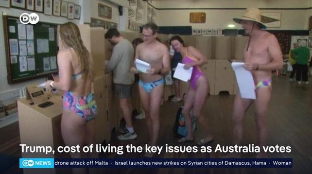 he image shows a group of people voting in a polling station. The individuals are dressed in swimwear, with some wearing colorful bathing suits and others in swimming trunks. They are standing in front of  voting booths, holding ballots. The setting appears to be indoors, with framed pictures on the walls and a bulletin board in the background. The floor is wooden, and there are other people visible in the background, some of whom are also in swimwear. The text overlay at the bottom of the image reads, "Trump, cost of living the key issues as Australia votes," along with additional news headlines about a drone attack off Malta, Israel's strikes on Syrian cities, and a woman.

Provided by @altbot, edited by @jemmesedi