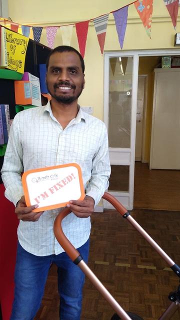A man with a pram frame holding a sign declaring that the item has been fixed.