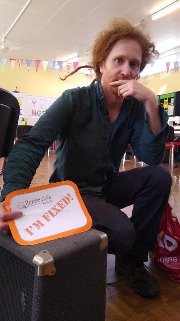 A man with a guitar amp holding a sign declaring that the item has been fixed.