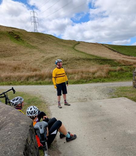 Two cyclists sit, resting by a wall, on the left. Just further on is another stood looking to the right while on a tarmac track. In the background are hills and a partially cloudy sky.