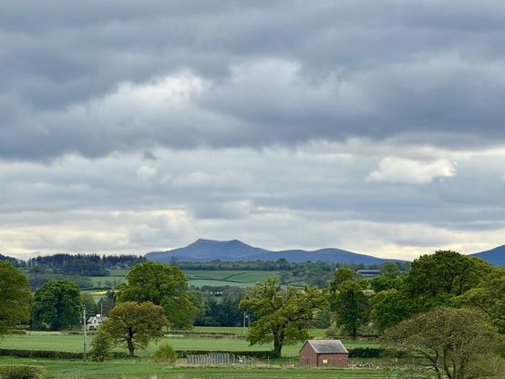A rural scene. Looking across green fields, hedgerows and leafy trees to a range of blue-grey mountains in the far distance. The highest has a dip in the middle making it look a little like a saddle. There is a small red brick building with a grey pitched roof in the lower right-hand part of the photo. The sky is a moody mix of grey and white cloud.