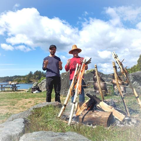 Two men stand on a grassy shoreline next to a campfire with salmon roasting. The man on the left has a beard and is wearing a blue shirt and black pants. The man on the right is wearing a cedar bark woven hat and a red long sleeve shirt with a native cedar tree design. Both men have their hands raised forward, palms facing outward. Over the fire are sticks propped up like a teepee, with salmon skewered and roasting. In the background is a blue sky with fluffy white clouds and a glimpse of the ocean.