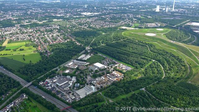 An aerial photo of the spoil heap Hoheward with the Horizon Observatory. There are trees all around. Part of a freeway can be seen at the bottom left. Multiple living quaters can be seen in the top left. There is a big factory in the top right.