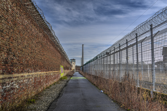 Foto eines Wachganges zwischen Mauer und Innenzaun eines ehemaligen Gefängnisses. im Hintergrund ein Wachtum mit teil zerschlagenen Fensterscheiben, Graffiti auf einem Teil der Mauer, Stacheldraht auf Zaun und Mauer.