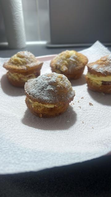 Four small cakes are placed on a paper towel. They appear to be mini sponge cakes or muffins, each split in half and filled with a cream or custard. The tops are dusted with powdered sugar. The pastries are arranged in a casual manner, and the lighting suggests they are in a kitchen setting.