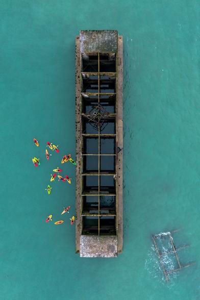 A vertical view of a concrete structure in the see. These were the floating docks, part of the "Mulberry B" harbour. A "flock" of kayakists are on the left of it.
