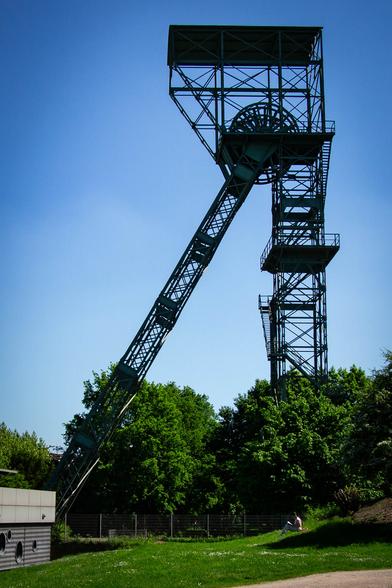 Color photo of an old coal mine headframe. The surroundings of the structure are now a park with some trees and grass areeas. A woman is reading on a lawn.