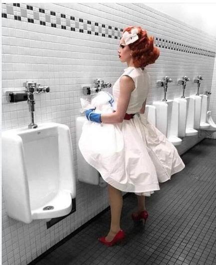In diesem Bild steht eine Person in einem weiß-rot akzentuierten 1950er-Jahre-Kleid mit Handschuhen, High Heels und gestylter roter Hochsteckfrisur vor einem Pissoir in einem klassischen Herrentoilettenraum. Die Szene wirkt bewusst inszeniert und spielt provokant mit Geschlechterrollen, öffentlichem Raum und der Wahrnehmung von Identität – eine visuelle Herausforderung an binäre Normen und gesellschaftliche Erwartungshaltungen.🖖