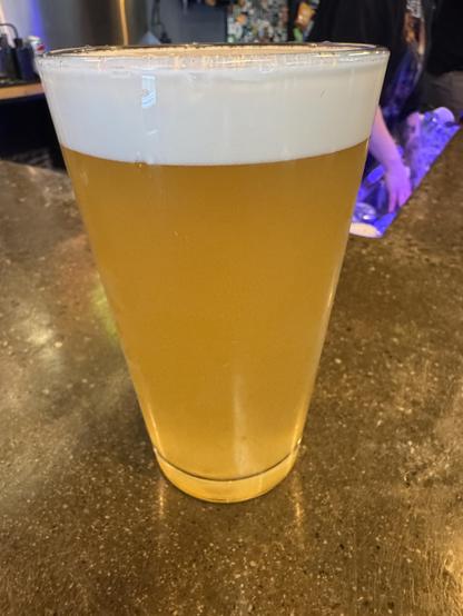 A pint of golden beer with a thick, frothy white head, sitting on a dark countertop.