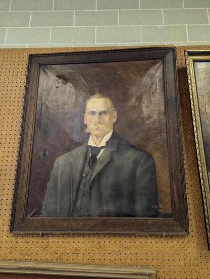 A painted portrait of a white man with a large bushy mustache in a black suit and tie, from the early 1900s. It is framed and hanging on a pegboard wall.