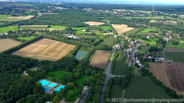 An aerial view of farm yards in Recklinghausen. Theres a street going through the image. A swimming pool sourrounded by trees is in the bottom left. There are some houses around the image.