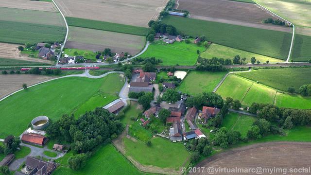 An aerial view of the countryside of Recklinghausen with some houses in between sourrounded by trees. A railway track is going through the image. A red train is driving on the track.