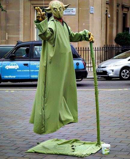 This image features a street performer dressed as Yoda from "Star Wars", performing a levitation illusion on a sidewalk. Yoda appears to be floating above the ground with the support of a single vertical rod covered in green material, which matches his robe, creating an interesting visual effect. There is a tip jar placed in front of him on the green cloth that is laid on the pavement. In the background, there are cars and a taxi with an advertisement on it, suggesting this performance is taking place in a city area, near Trafalgar Square in London according to the sign seen behind.