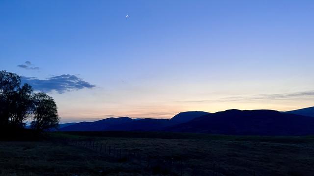The Monadhliath Mountains silhouetted against the last glimmer of the setting sun. The foreground is very dark green, with bluish mountains behind. The sky fades from dusky blue to pale orange, and the new moon is visible at the top.