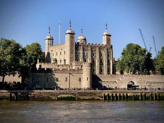 This image shows the Tower of London, a historic fortress and former royal palace in London, England. The photo captures a view of the tower beside the River Thames. You can see the distinct medieval architecture with the stone walls and turret-style towers topped with four spires each. The entrance called "Traitor's Gate" is visible at riverside level, historically used to bring prisoners by boat into the tower. The scene includes blue skies and some greenery around the structure, with a few people visible along the riverside walkway.