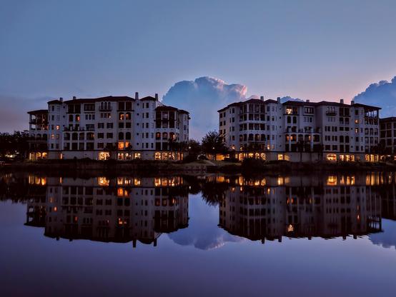 Beautiful blue hour view across a quiet waterway where large luxurious buildings on the opposite shoreline cast their shadows and light reflections upon the water below, while large blue cloud formations rise behind them, illuminated by shades of pink and purple from the approaching dawn.