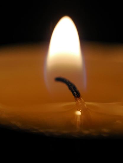 Nahaufnahme einer Kerzenflamme. Die Kerze ist gelbbraun, der Docht ist in der Mitte des Bildes nach links geneigt. Der Hintergrund ist schwarz.