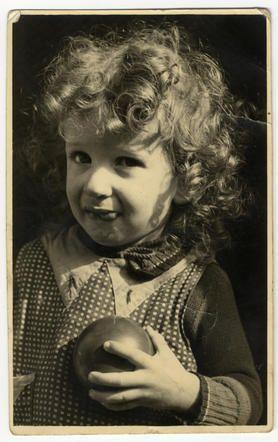 A vintage black and white photo of a young girl with curly hair, holding an apple and smiling slightly.