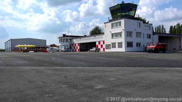 A photo of the main hanger and tower of the Marl Lohemühle airport. There is a sports planes in front of the hangar and a fire truck right of it. Next to the tower building on the left is a small open-air restaurant.