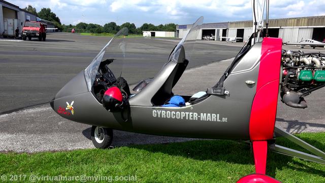A photo of the gyrocopter from the side, parking on a grass area near the hangars. There are some sports planes in front of the hangars and a helicopter in the back. A fire truck is standing next to a hangar.