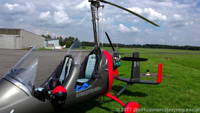 A photo of the gyrocopter from the front side, parking on a grass area near the hangars. The runway can be seen at the back.