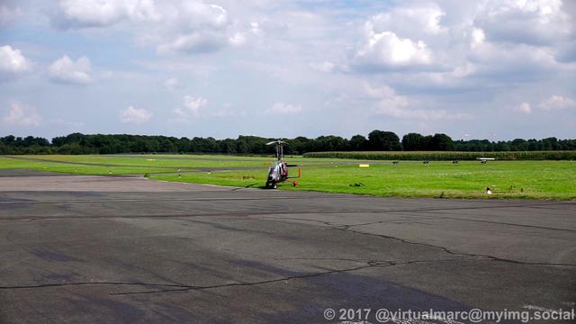 A photo of the gyrocopter from the front, parking on a grass area near the hangars. The runway can be seen at the back.