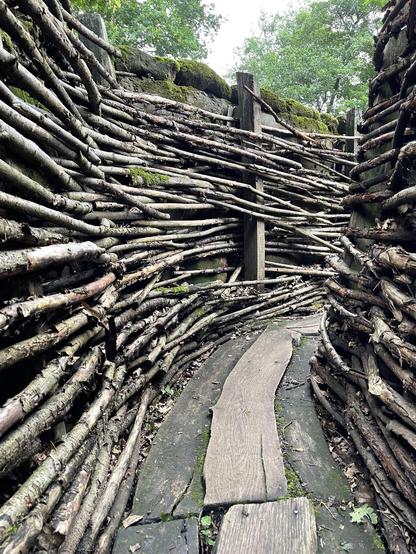 The image you provided shows a wooden path crafted from rough, interwoven branches and logs. This rustic pathway meanders through an outdoor area, intended to facilitate walking across potentially uneven or muddy terrain. Surrounding the pathway are vegetation and large stones, suggesting that this place could be a forested or a natural, potentially preserved area designed for walking and experiencing nature. The use of natural materials blends the path nicely with the surrounding environment, enhancing the ecological aesthetics and minimizing the impact of the trail on the natural setting.