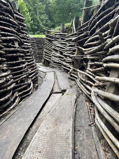 The image shows a wooden pathway lined with walls made from woven branches and sticks. This pathway is constructed with split logs laid out as flat surfaces to walk on, and the surrounding barriers appear to be built using a rustic, wickerwork technique with naturally shaped wood. The scene looks like it might be set in a forested or garden area given the lush green trees in the background. This kind of pathway could be part of a nature trail, garden, or an outdoor historical or cultural exhibit.