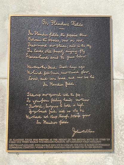 This image shows a plaque displaying the famous poem "In Flanders Fields" written by John McCrae. The plaque has embossed gold lettering on a dark background. Below the poem, there's additional text noting historical context:
- The poem was written during the height of the Second Battle of Ypres on 3rd May 1915.
- John McCrae was a brigade-surgeon of the 1st Canadian Field Artillery.
- McCrae died on 28th January 1918 and is buried in Wimereux Communal Cemetery, where a stone memorial seat is erected in his memory.

The poem itself articulates the experience of World War I and expresses mourning for the fallen soldiers, symbolized through the image of poppies growing between crosses marking the graves.