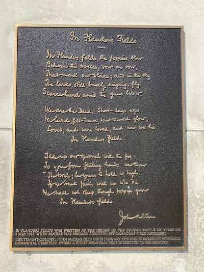 This image shows a plaque displaying the famous poem "In Flanders Fields" written by John McCrae. The plaque has embossed gold lettering on a dark background. Below the poem, there's additional text noting historical context:
- The poem was written during the height of the Second Battle of Ypres on 3rd May 1915.
- John McCrae was a brigade-surgeon of the 1st Canadian Field Artillery.
- McCrae died on 28th January 1918 and is buried in Wimereux Communal Cemetery, where a stone memorial seat is erected in his memory.

The poem itself articulates the experience of World War I and expresses mourning for the fallen soldiers, symbolized through the image of poppies growing between crosses marking the graves.