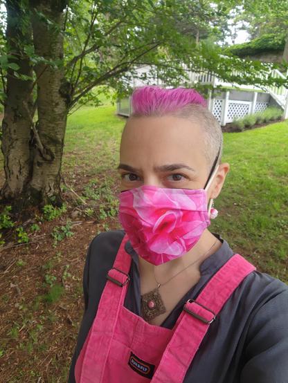 Selfie, in a very green park setting. I'm wearing pink velvet dungarees over a loose grey shirt with a small standing collar. Pendant is a silver filigree square, hanging by one of the corners, with a small round pink stone in the center. Dangling earrings are small light pink peonies. Face mask has a bright pink dahlia close-up print. Hair also pink, of course.