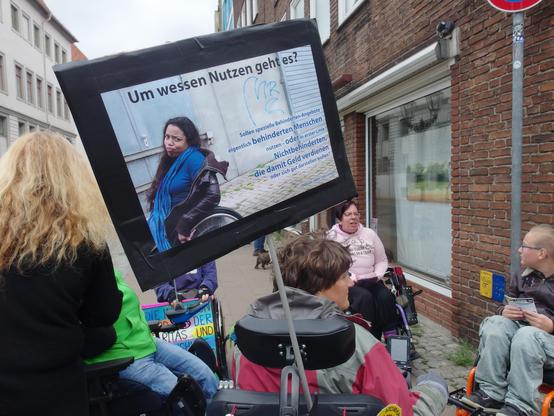 Demonstrierende, ein Plakat: Eine junge, afro-deutsche Frau fährt mit ihrem Rollstuhl seitlich aus dem Bild heraus. Sie guckt verärgert in die Kamera und zieht eine Grimasse.
Ihr Text: Um wessen Nutzen geht es? Sollen spezielle Behinderten-Angebote eigentlich behinderten Menschen nutzen – oder in erster Linie Nichtbehinderten, die damit Geld verdienen oder sich gut darstellen wollen? 
Zur Person: Désirée, mehr Infos online