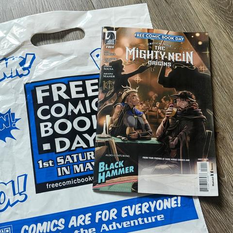 A white bag with blue "FREE COMIC BOOK DAY" text has a comic book on it. The comic is "Critical Role: THE MIGHTY NEIN ORIGINS" and also features "BLACK HAMMER".